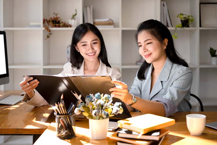 Two Asian women sitting at office desk and talking about marketing ideas.