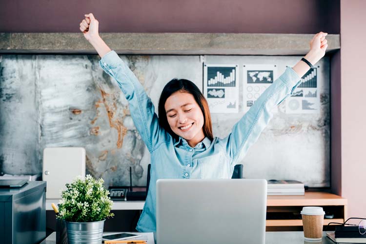 Woman raises arms with closed eyes in front of a laptop on the table.