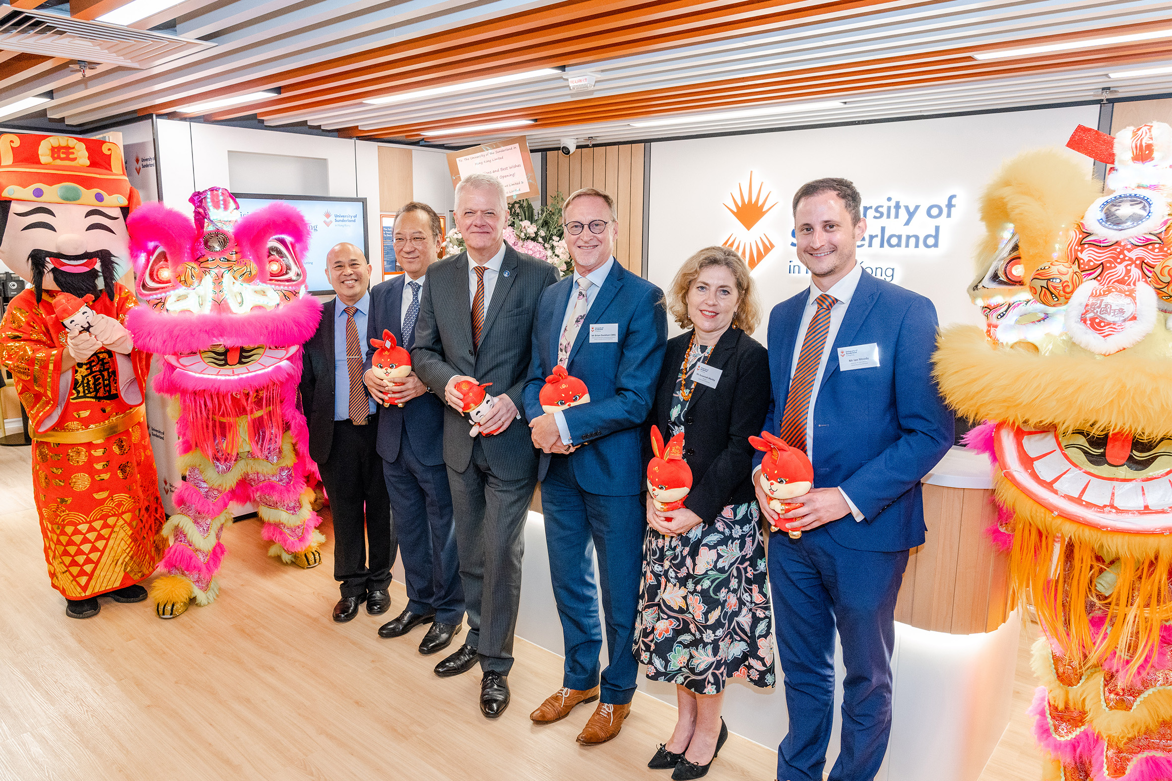 A group of performers dressed in traditional lion dance costumes, performing at the opening ceremony of a new campus. They are surrounded by a crowd of people, including University of Sunderland staff and guests of honour, who are watching the performance. The background is the reception of University of Sunderland in Hong Kong's new campus.