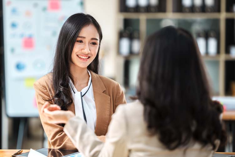 Asian businesswoman holding cup of coffee, smiling and looking at camera while sitting at office desk.