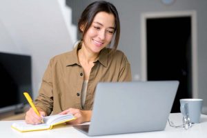 Smiling young woman having an online lesson in front of the laptop and notebook.
