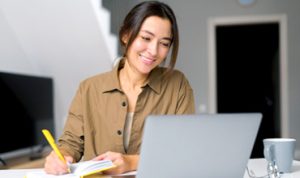 Smiling young woman having an online lesson in front of the laptop and notebook.