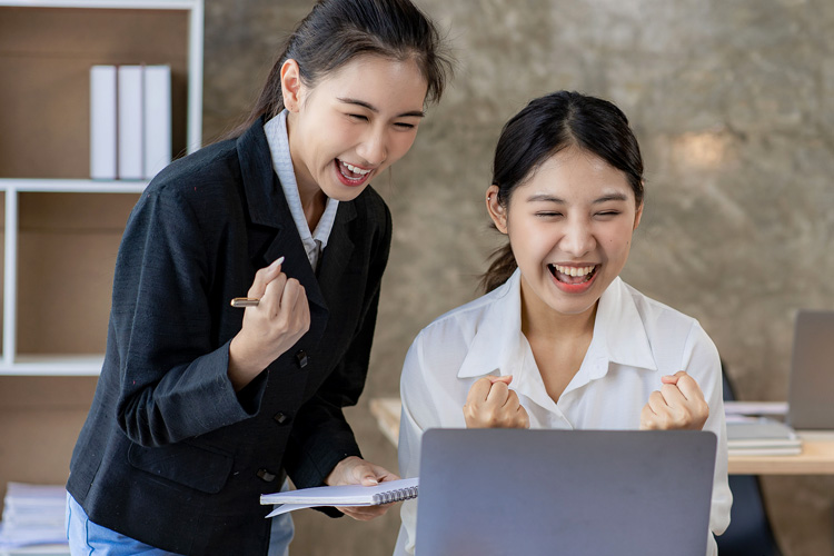 Two Asian women celebrate business success at the office with laptops and financial graphs.