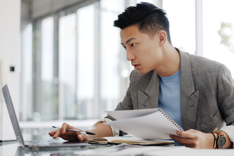 Asian man working on laptop with papers, focused in office setting.