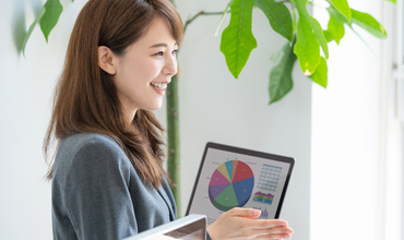 Asian woman presenting financial data on a laptop, smiling, in office setting.