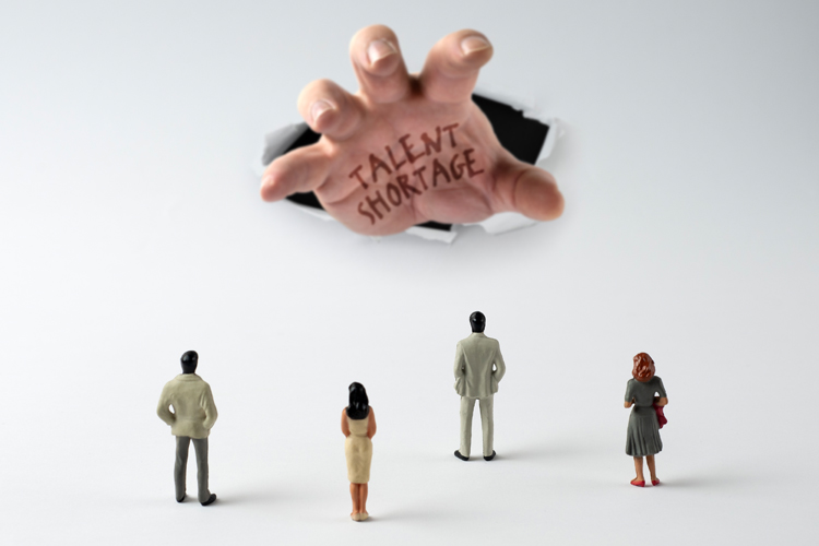 People look up at a large hand emerging with "Talent Shortage" written on it.
