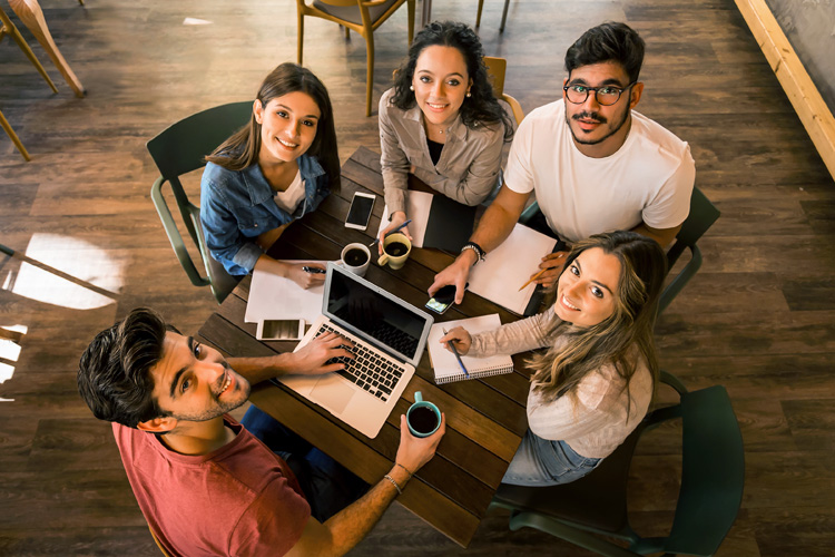 Five young students studying together at a table with laptops and coffee.