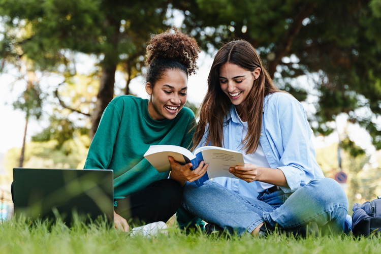 Two students sitting on public park grass learning together with book and laptop.