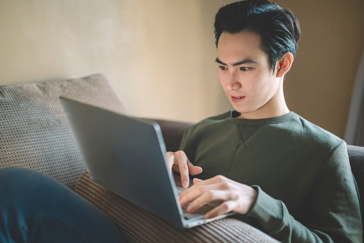 A young man sitting on a couch, focused on his laptop, wearing a green sweatshirt