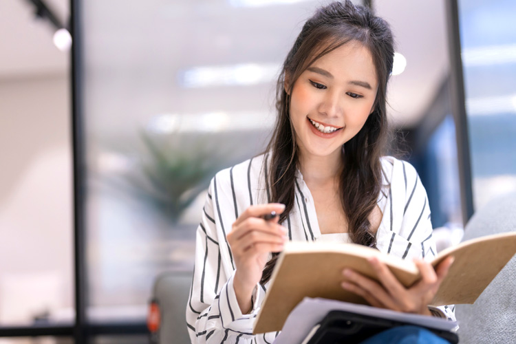 A smart casual Asian woman sits joyfully in her living area, jotting ideas in a notebook.
