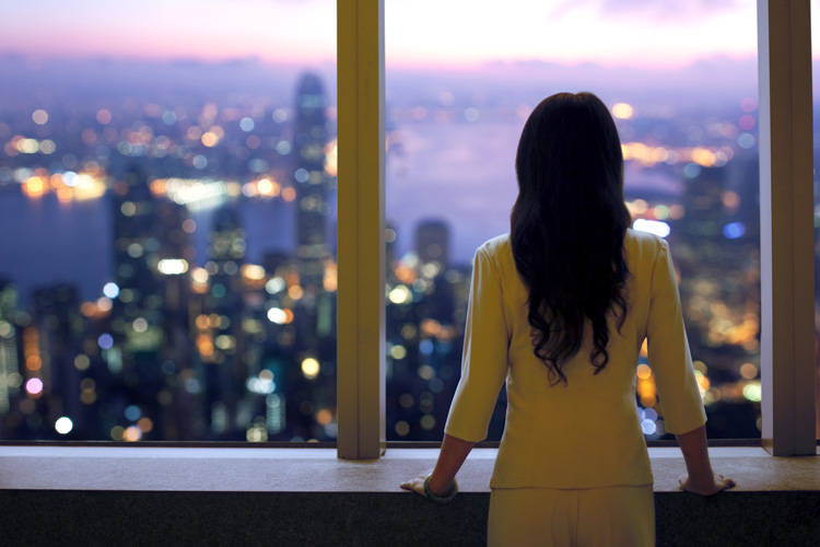 A young woman in a suit gazes at the Hong Kong skyline from a building window.