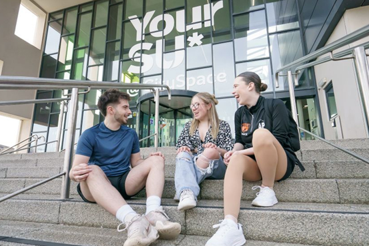 Three students sitting on the steps outside the Your SU building at the University of Sunderland.