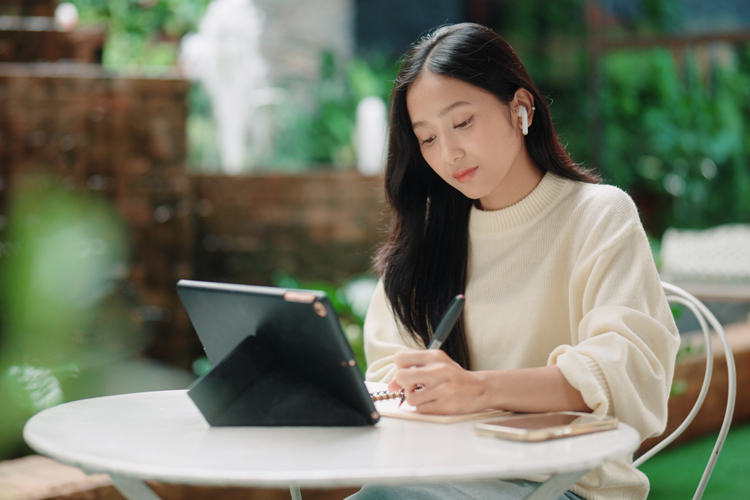 Focused female student writing notes in a notebook during an online lesson on her digital tablet.