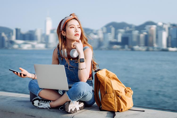Thoughtful Asian woman in headphones, sitting cross-legged by the promenade, using smartphone and laptop.