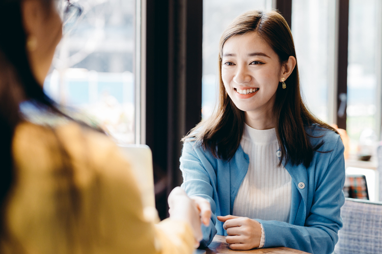 A smiling young woman shakes hands with another person in a cozy cafe setting.