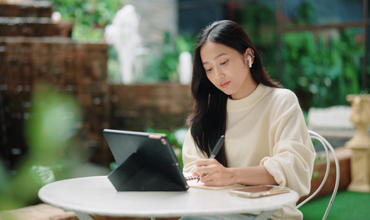 Focused female student writing notes in a notebook during an online lesson on her digital tablet.